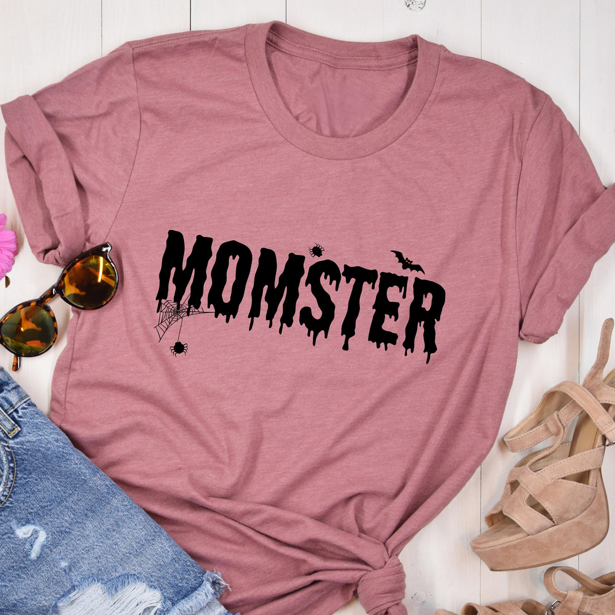 May include: A dusty rose t-shirt with the word "MOMSTER" in black, dripping font. A spider web and bat graphic are above the word. The shirt is folded, with sunglasses and denim shorts visible.