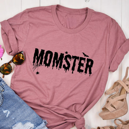 May include: A dusty rose t-shirt with the word "MOMSTER" in black, dripping font. A spider web and bat graphic are above the word. The shirt is folded, with sunglasses and denim shorts visible.