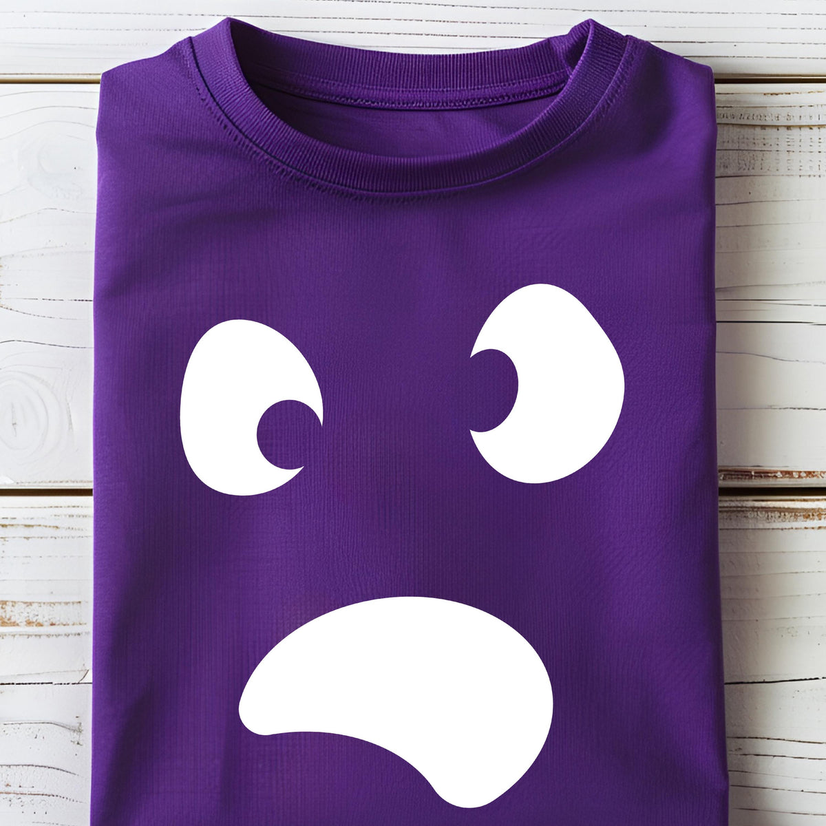 May include: A folded purple t-shirt featuring a white cartoon face design. The design includes two crescent-shaped eyes and a curved mouth, creating a sad expression. The shirt is made of a soft material.