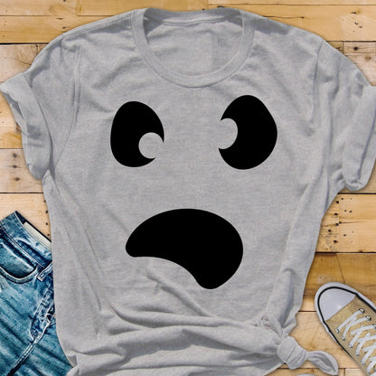 May include: A light gray t-shirt featuring a large, black ghost face design. The ghost face has two curved eyes and a wide, open mouth. The shirt is made of a soft material and is suitable for Halloween.