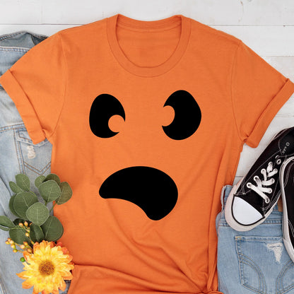 May include: Orange t-shirt with a black, surprised-face graphic. The graphic features two curved eye shapes and a curved mouth. The shirt is folded at the sleeves. The shirt is on top of a denim jacket, a pair of black sneakers, and a bouquet of flowers.
