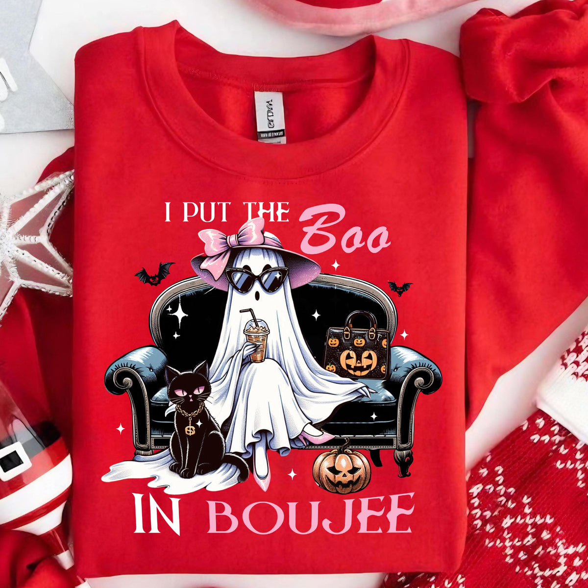 May include: A bright red crewneck sweatshirt featuring a graphic of a ghost wearing a pink bow and sunglasses, sitting on a black sofa. The ghost holds a coffee cup next to a black cat and a pumpkin. Text reads I PUT THE Boo IN BOUJEE.