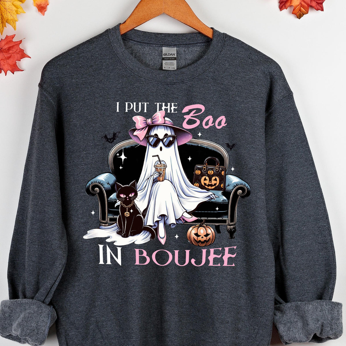May include: A dark heather gray crewneck sweatshirt featuring a graphic of a ghost wearing a pink hat and sunglasses, sitting on a black sofa with a black cat, a pumpkin, and a handbag. The text reads I PUT THE BOO IN BOUJEE in pink letters.