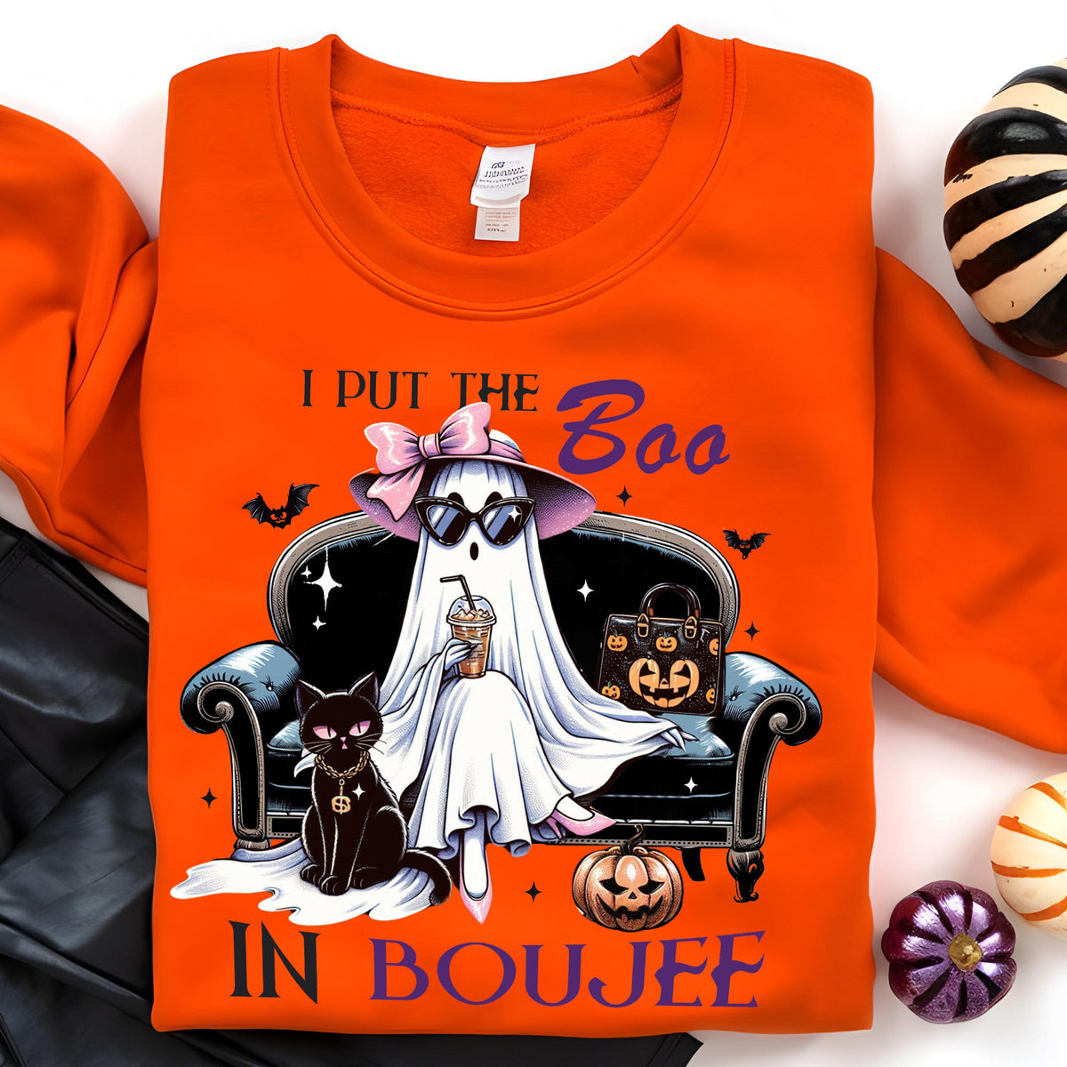 May include: An orange sweatshirt featuring a graphic of a ghost wearing a pink bow, sunglasses, and holding a coffee cup while sitting on a sofa next to a black cat and a pumpkin-patterned handbag, with the text I PUT THE BOO IN BOUJEE.