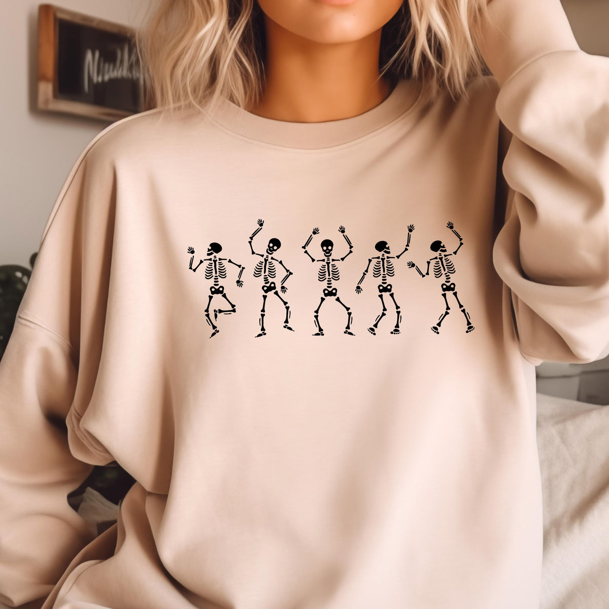 May include: Beige sweatshirt featuring a black graphic of five dancing skeletons. The skeletons are in various poses with their arms raised. The sweatshirt has a crew neck and long sleeves. The overall theme is Halloween.