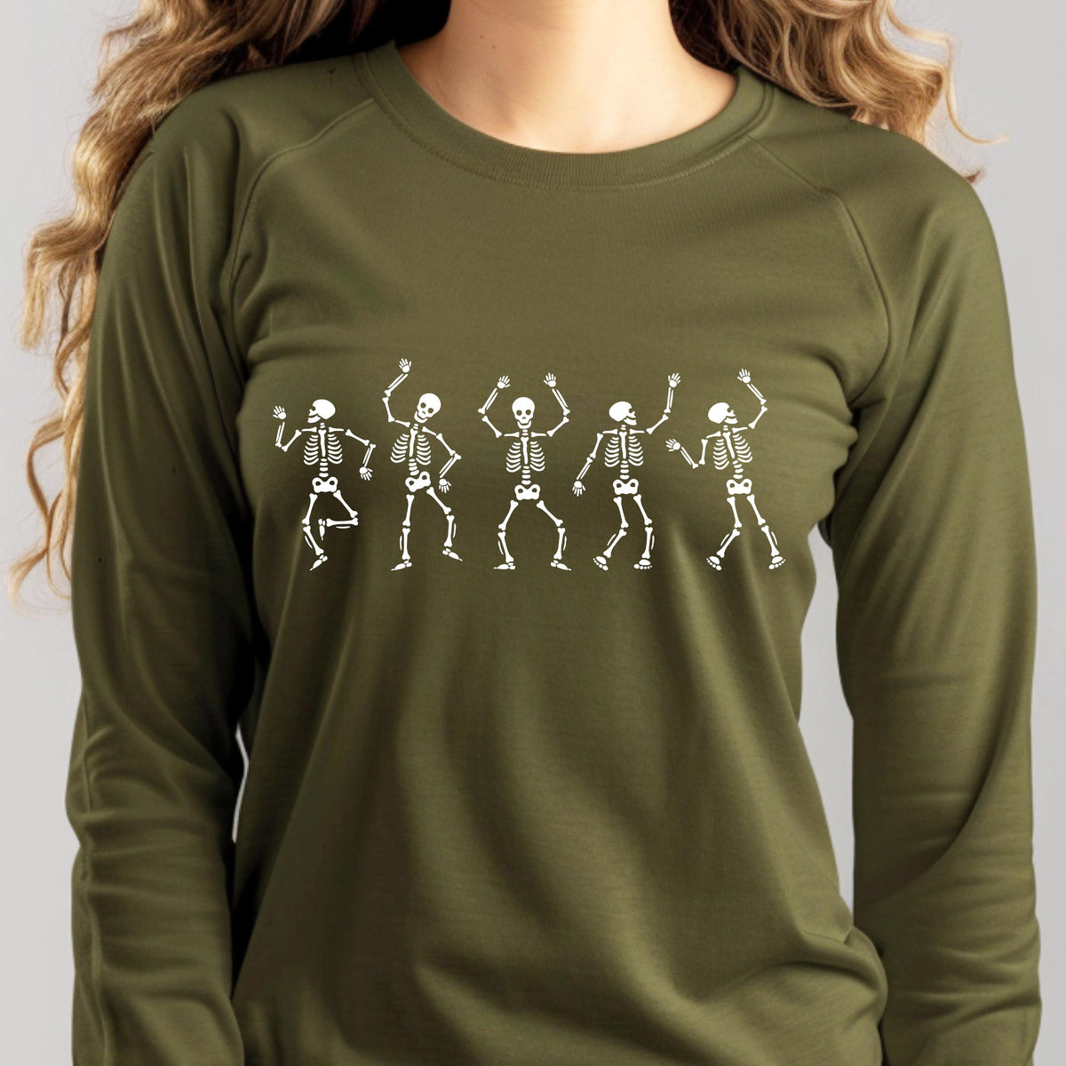 May include: Olive green long-sleeve shirt featuring a white graphic of five dancing skeletons. The skeletons are in various poses, with their arms raised and legs bent, creating a playful and festive design. The shirt is made of a soft material.
