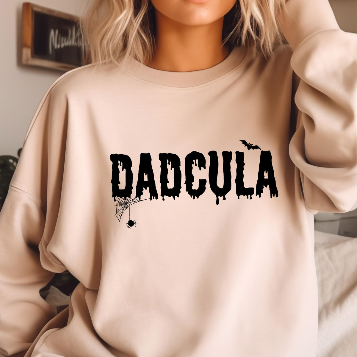 May include: A light beige sweatshirt with a black graphic print that says "DADCULA" with a dripping effect. A spider web and spider are below the text and a bat is above the text.