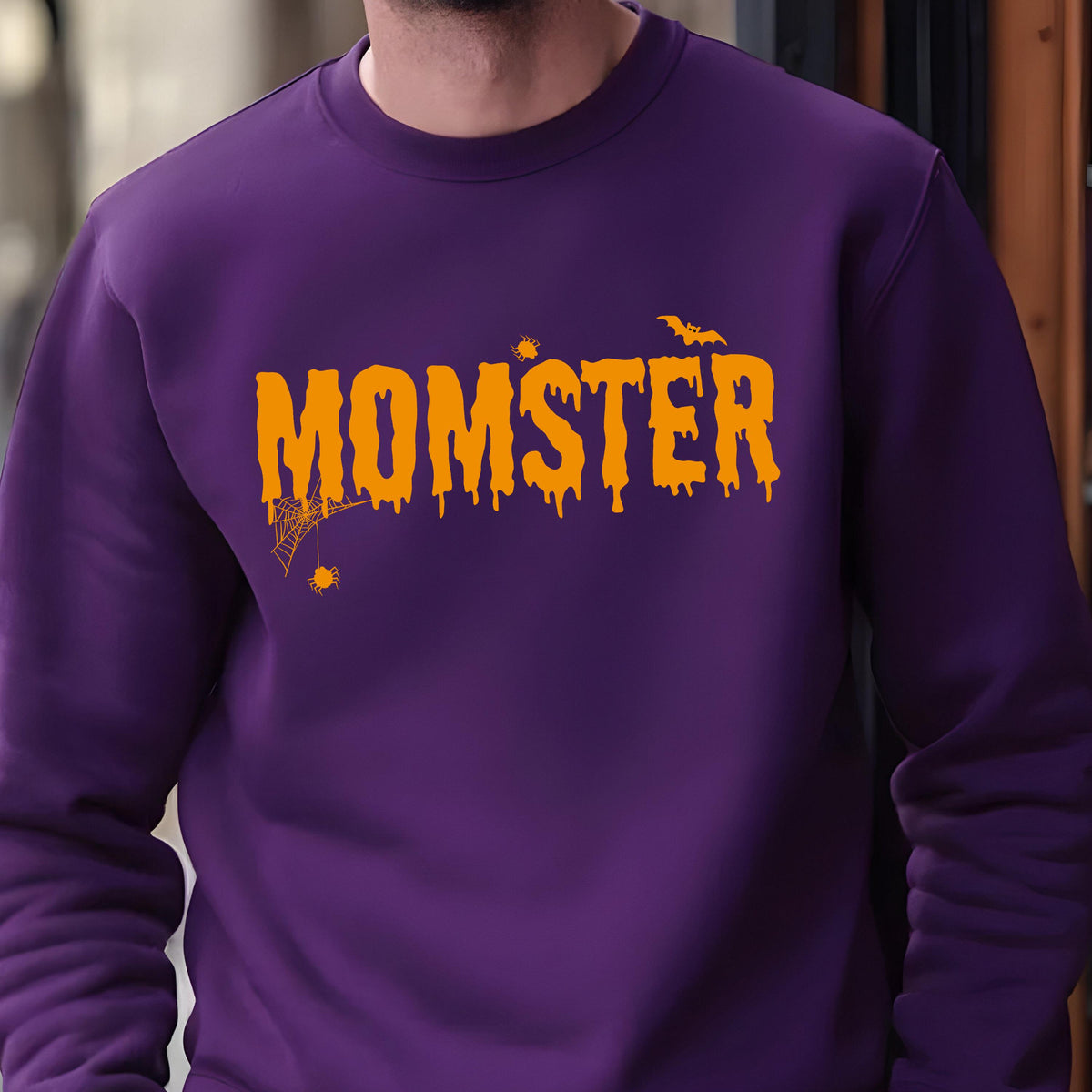 May include: A purple long-sleeve crewneck sweatshirt featuring the orange text MOMSTER in a dripping font. The design includes a small spider hanging from a web, a tiny spider, and a flying bat silhouette positioned above the lettering.