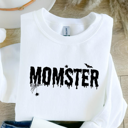 May include: A white long-sleeve sweatshirt featuring the black text MOMSTER in a dripping font. The design includes a spider hanging from a web on the letter M, a small spider above the letter S, and a flying bat silhouette above the letter T.