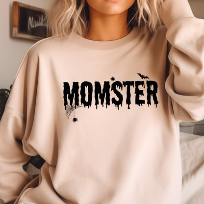 May include: A beige crewneck sweatshirt featuring the black text MOMSTER in a dripping font style. The design includes a small spider web with a hanging spider on the left side, a tiny spider above the letter T, and a flying bat above the letter R.