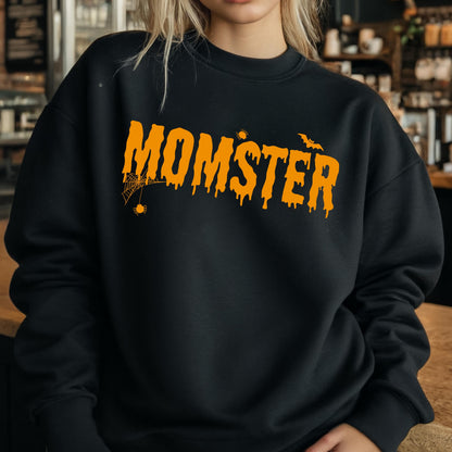 May include: A black crewneck sweatshirt featuring the orange text MOMSTER in a dripping font style. The design includes a small spider hanging from a web on the letter M, a tiny spider above the letter S, and a small bat flying near the letter R.