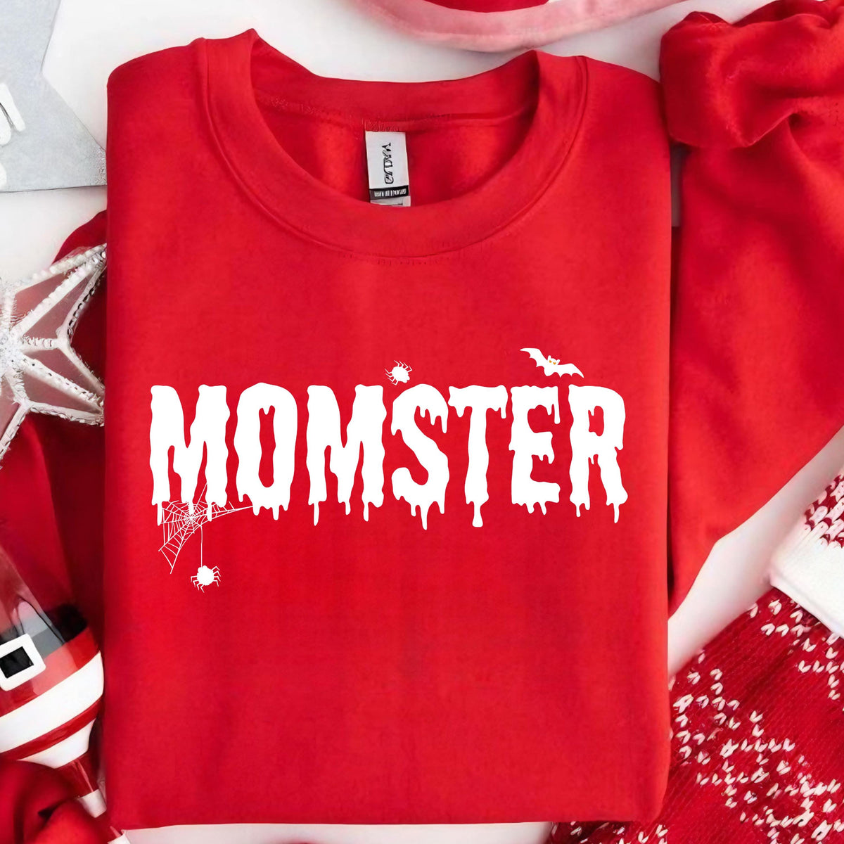 May include: A bright red crewneck sweatshirt featuring the white text MOMSTER in a dripping, spooky font. The design includes a small spider web with a hanging spider, a tiny spider, and a bat silhouette integrated into the lettering.