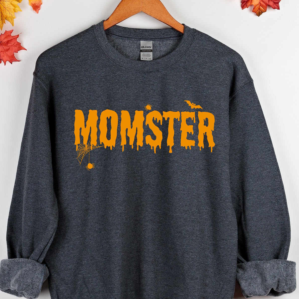 May include: A dark heather gray long-sleeve crewneck sweatshirt featuring the orange text MOMSTER in a dripping font. The design includes a small orange spider hanging from a web on the letter M and a flying bat silhouette above the letter R.