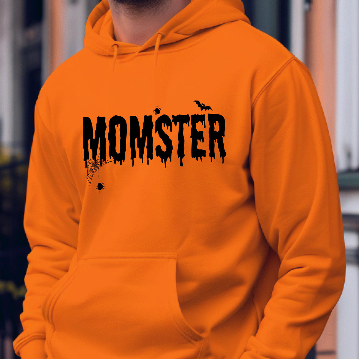 May include: A bright orange hooded sweatshirt featuring the black text MOMSTER in a dripping font style. The design includes a small spider web with a hanging spider on the left, a tiny spider above the text, and a small flying bat on the right.