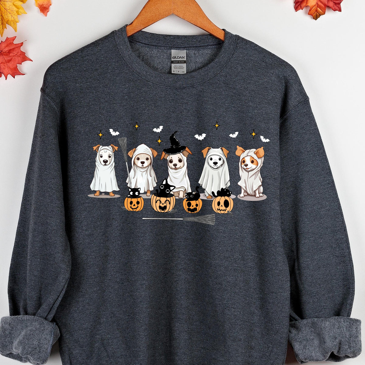 May include: Dark gray sweatshirt featuring a Halloween-themed graphic. The design includes five dogs dressed in costumes, including ghosts and a witch. Pumpkins, bats, and a broom are also part of the illustration. The sweatshirt is hanging on a wooden hanger.