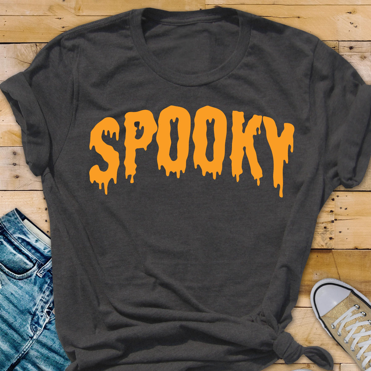 May include: Dark gray t-shirt with the word "SPOOKY" in orange, dripping letters. The shirt is a crew neck style and is knotted at the bottom. The shirt is on a wooden surface with a pair of jeans and a sneaker.