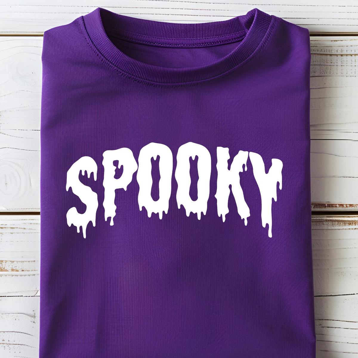 May include: A folded purple t-shirt with the word "SPOOKY" in dripping white letters. The shirt is made of a soft material and is likely for Halloween or spooky-themed events.
