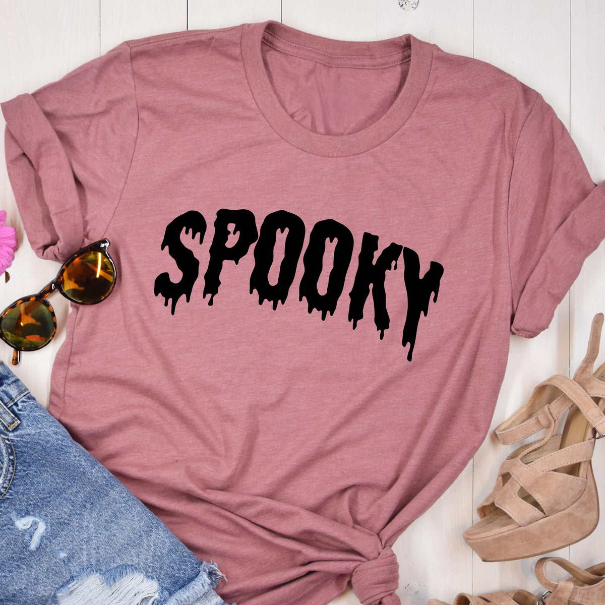 May include: A dusty rose-colored t-shirt with the word "SPOOKY" in black, dripping font. The shirt is a crew neck style with rolled-up sleeves. The image includes sunglasses, denim shorts, and platform sandals.