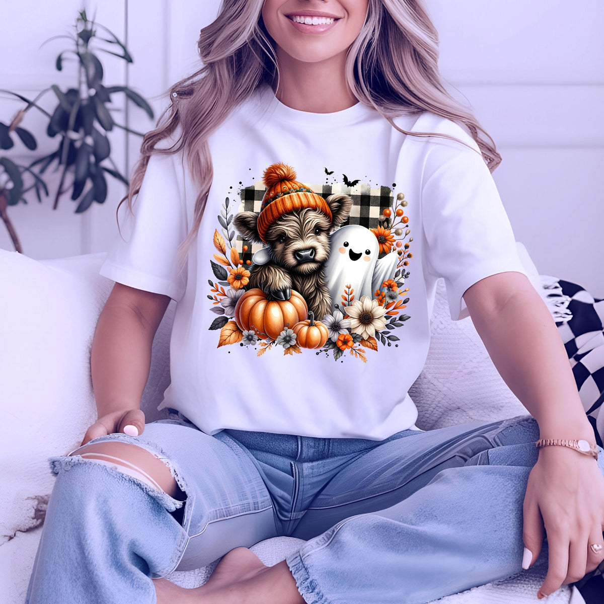 May include: A white t-shirt featuring a graphic print of a fluffy highland cow wearing an orange knit hat, accompanied by a small white ghost, orange pumpkins, autumn flowers, and a black and white buffalo plaid square background pattern.