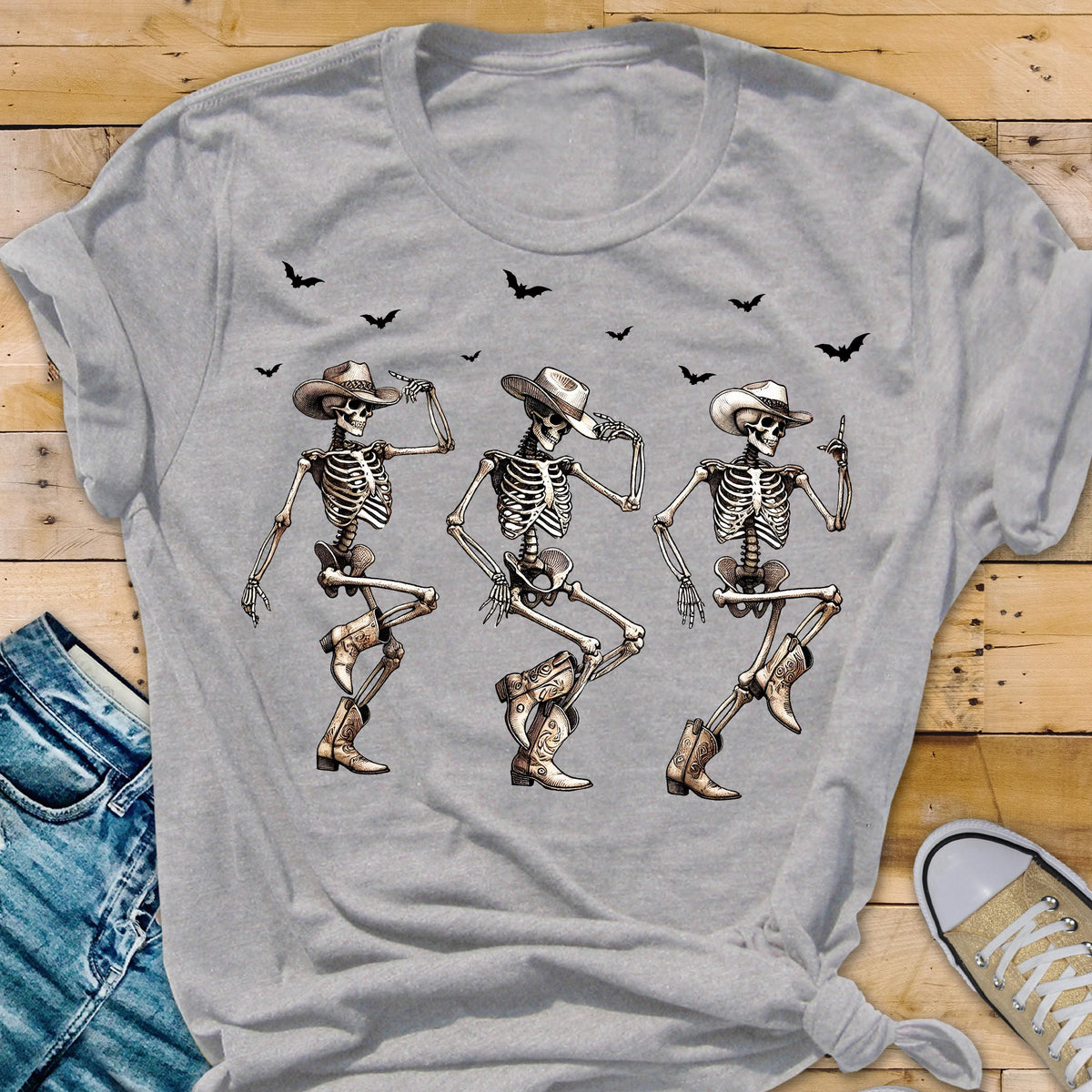 May include: A light gray t-shirt featuring a graphic of three dancing skeletons wearing cowboy hats and boots. Black bats are flying above the skeletons. The shirt is made of a soft material.
