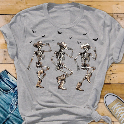 May include: A light gray t-shirt featuring a graphic of three dancing skeletons wearing cowboy hats and boots. Black bats are flying above the skeletons. The shirt is made of a soft material.