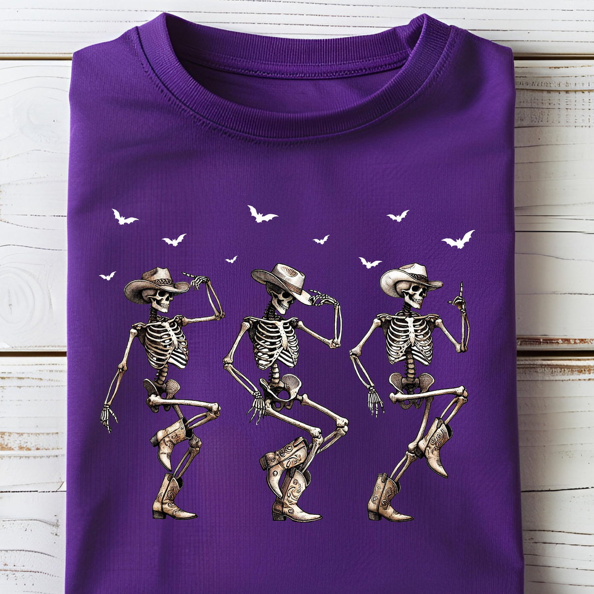 May include: A folded purple t-shirt featuring a graphic of three dancing skeletons wearing cowboy hats and boots. White bats are scattered across the top of the shirt. The skeletons are in a line, each in a different dance pose.