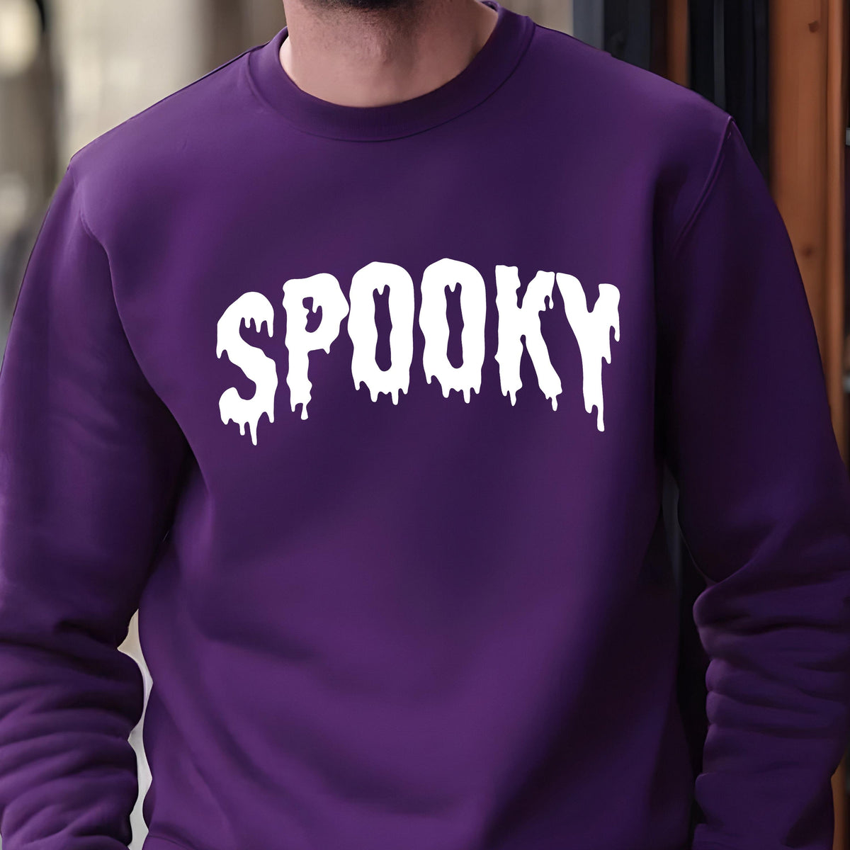 May include: A deep purple long-sleeve crewneck sweatshirt featuring the word SPOOKY printed in a white, dripping, liquid-style font across the chest. The garment has a ribbed neckline and cuffs, providing a clean and structured appearance.