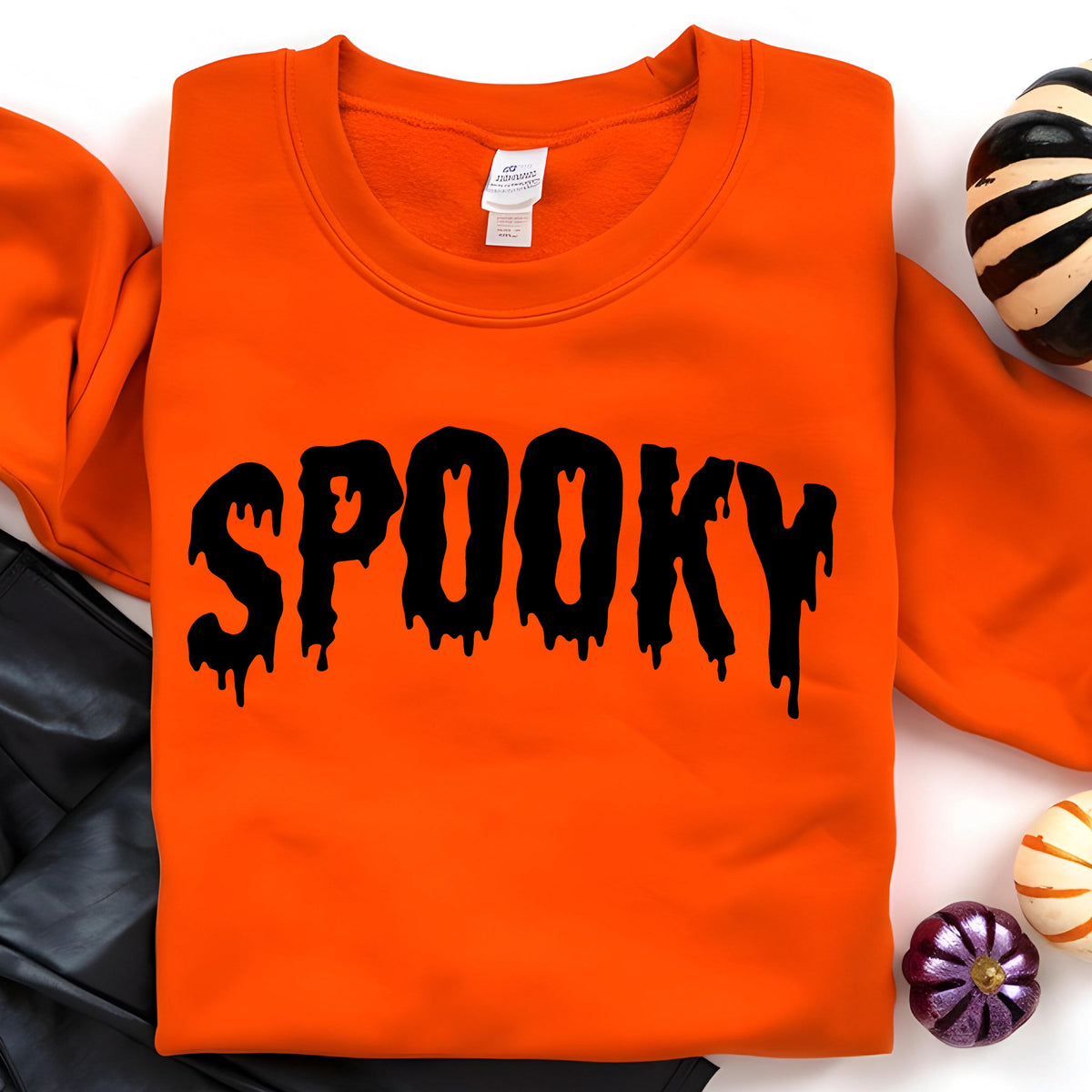 May include: A bright orange long-sleeve crewneck sweatshirt featuring the word SPOOKY printed in a black, dripping, liquid-style font across the chest. The garment has a standard ribbed neckline and a white interior brand label at the collar.