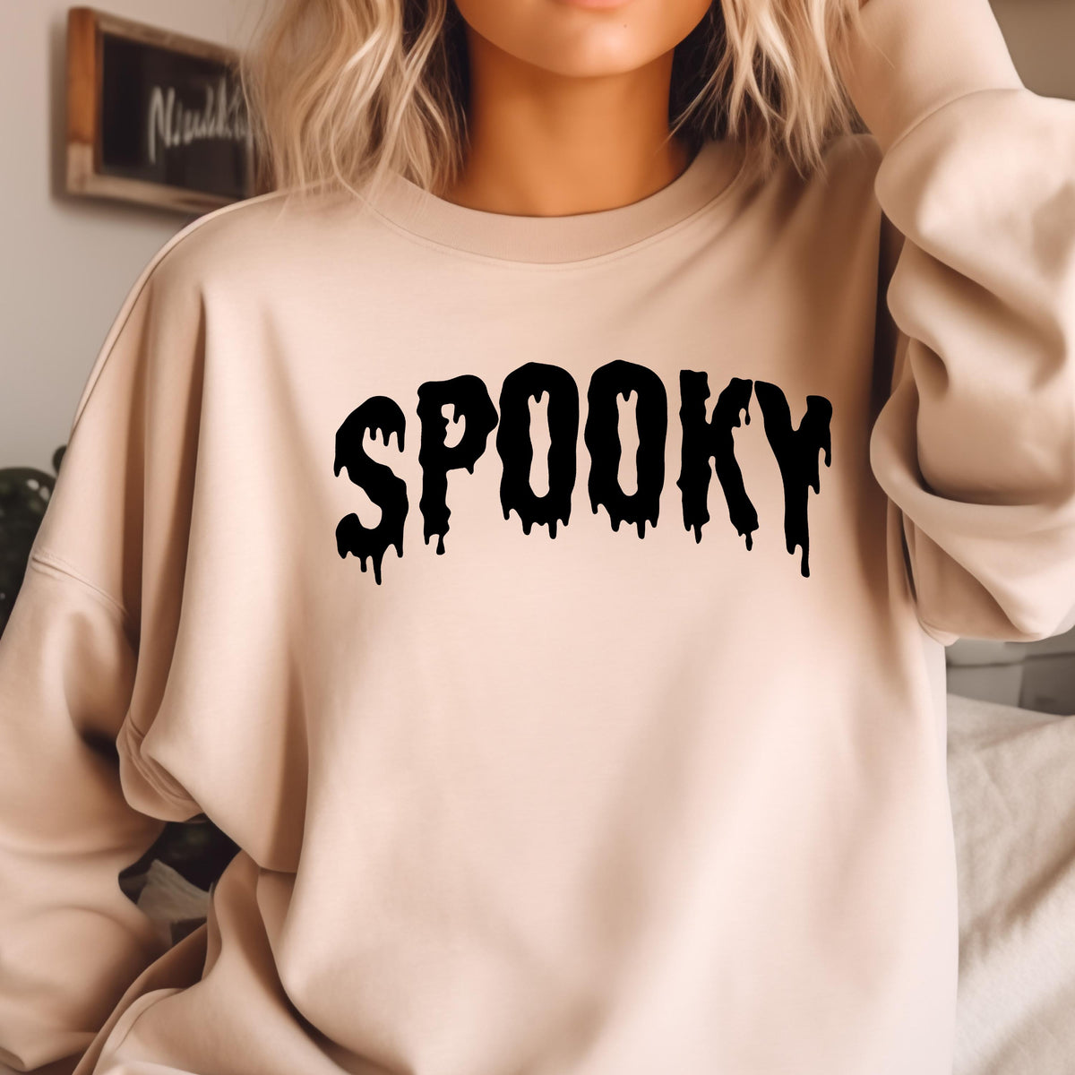May include: A beige crewneck sweatshirt featuring the word SPOOKY printed in a black, dripping, liquid-style font across the chest. The garment has long sleeves, a ribbed neckline, and a relaxed fit, providing a simple and clean aesthetic.