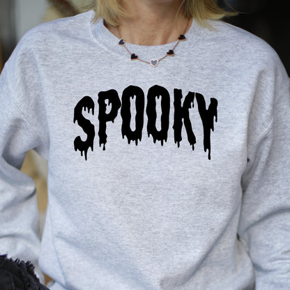 May include: A light gray heathered crewneck sweatshirt featuring the word SPOOKY printed in a black, dripping, liquid-style font across the chest. The garment has long sleeves and a ribbed neckline, paired with a delicate gold-tone heart necklace.