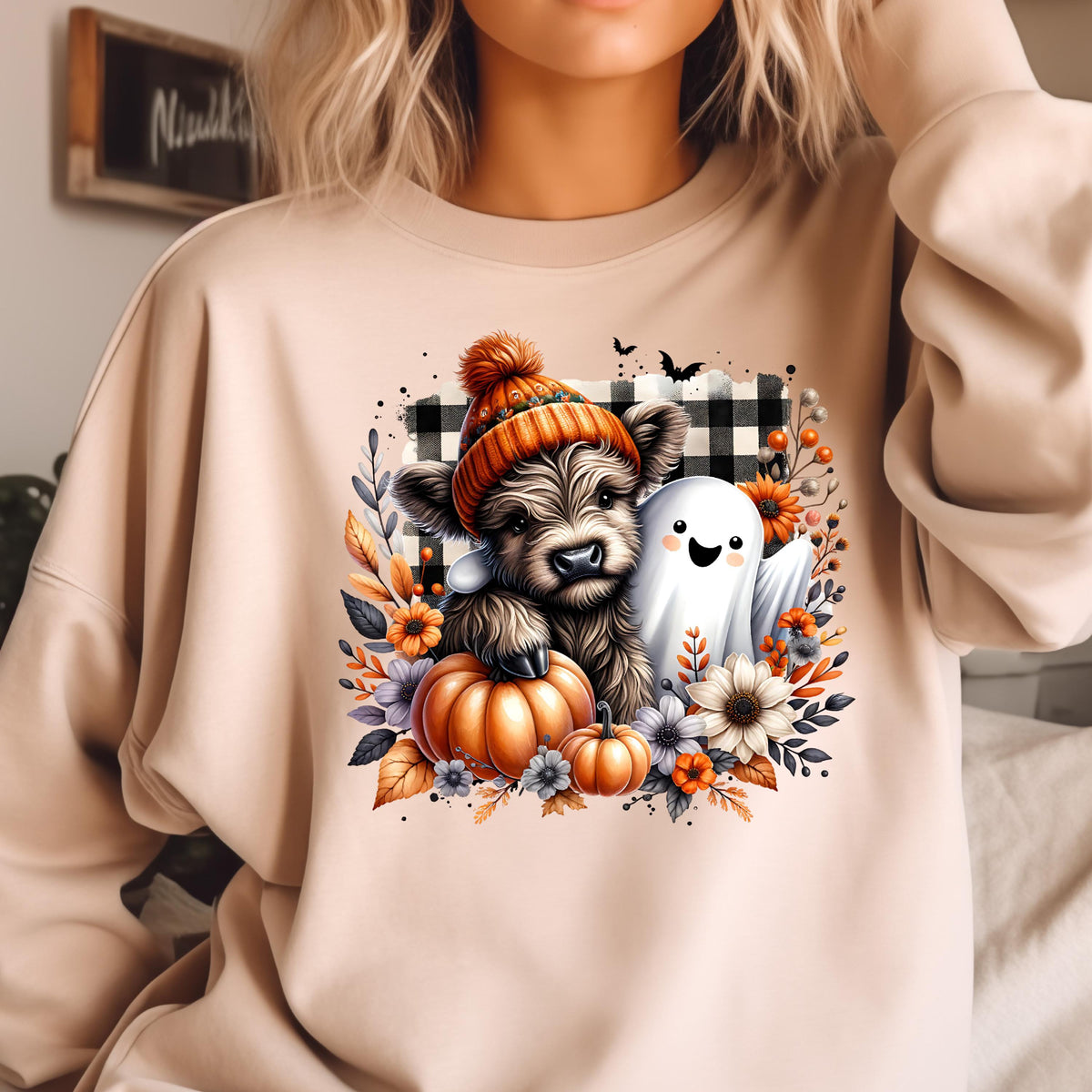 May include: A beige crewneck sweatshirt featuring a graphic print of a fluffy Highland cow wearing an orange knit hat, a small white ghost, orange pumpkins, and autumn flowers set against a black and white buffalo plaid square background.
