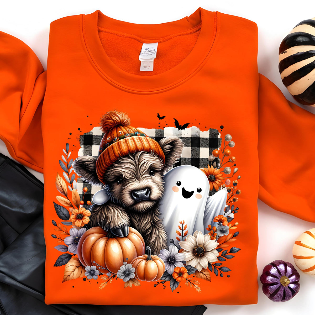 May include: An orange sweatshirt featuring a graphic of a fluffy highland cow wearing an orange knit hat, a white ghost, and pumpkins. The design is set against a black and white buffalo plaid background, surrounded by autumn flowers and leaves.