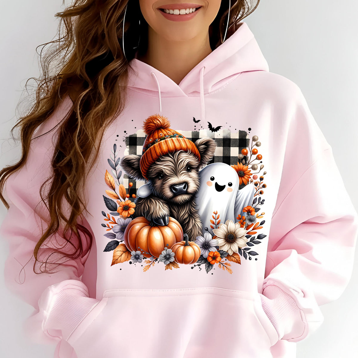 May include: A light pink hooded sweatshirt featuring a graphic print of a fluffy brown calf wearing an orange knit hat, a small white ghost, orange pumpkins, and autumn flowers set against a black and white buffalo plaid square background.