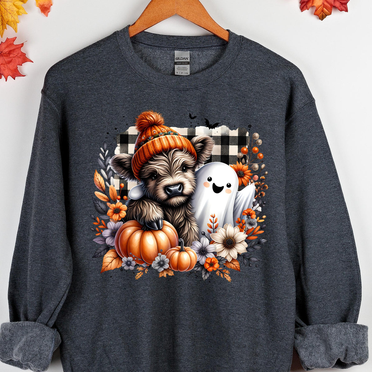 May include: A dark gray crewneck sweatshirt featuring a graphic of a fluffy highland cow wearing an orange knit beanie, standing next to a white ghost. The design includes orange pumpkins, autumn flowers, and a black and white buffalo plaid square.