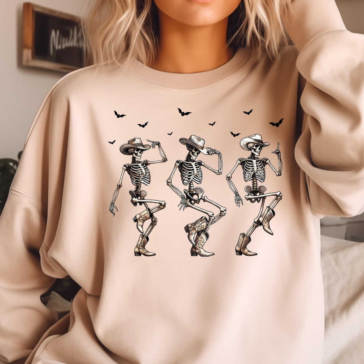 May include: A beige sweatshirt featuring a graphic of three dancing skeletons wearing cowboy hats and boots. Black bat silhouettes are scattered above the skeletons. The design is printed on a neutral-toned long-sleeved shirt.