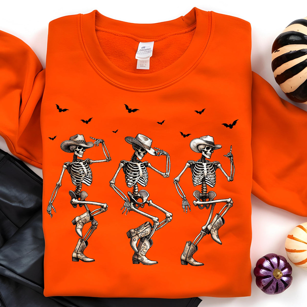 May include: Orange sweatshirt featuring a graphic of three dancing skeletons wearing cowboy hats and boots. Black bats are scattered across the top. The sweatshirt is a Halloween-themed apparel item.