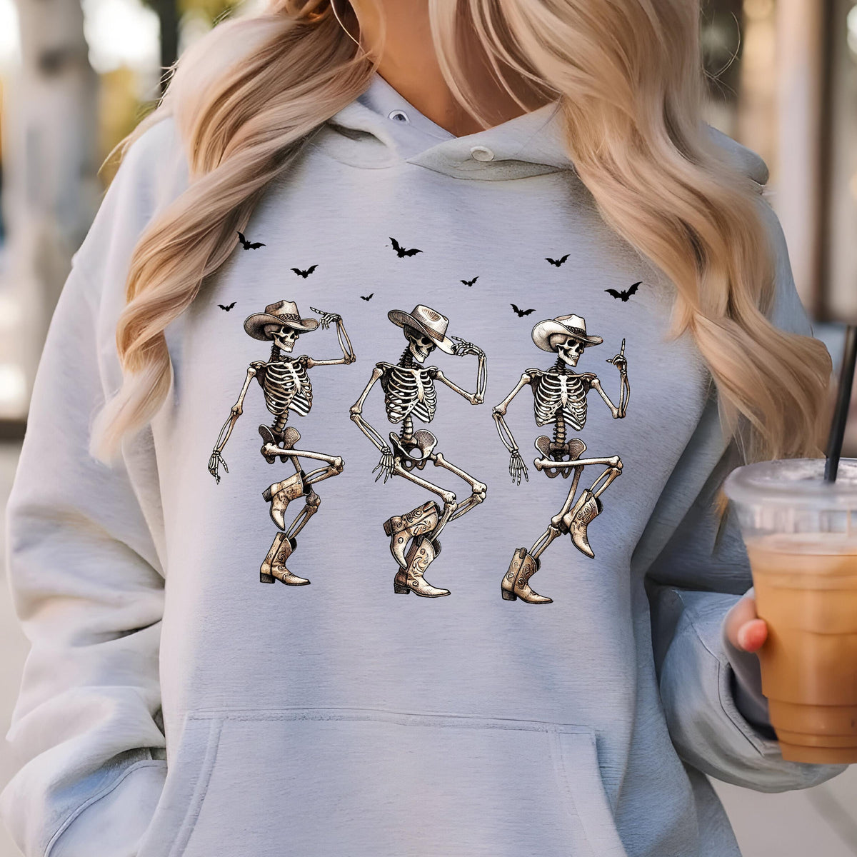 May include: Light blue hoodie featuring a graphic of three dancing skeletons wearing cowboy hats and boots. Black bats are flying above the skeletons. The design is a fun, festive theme for Halloween.