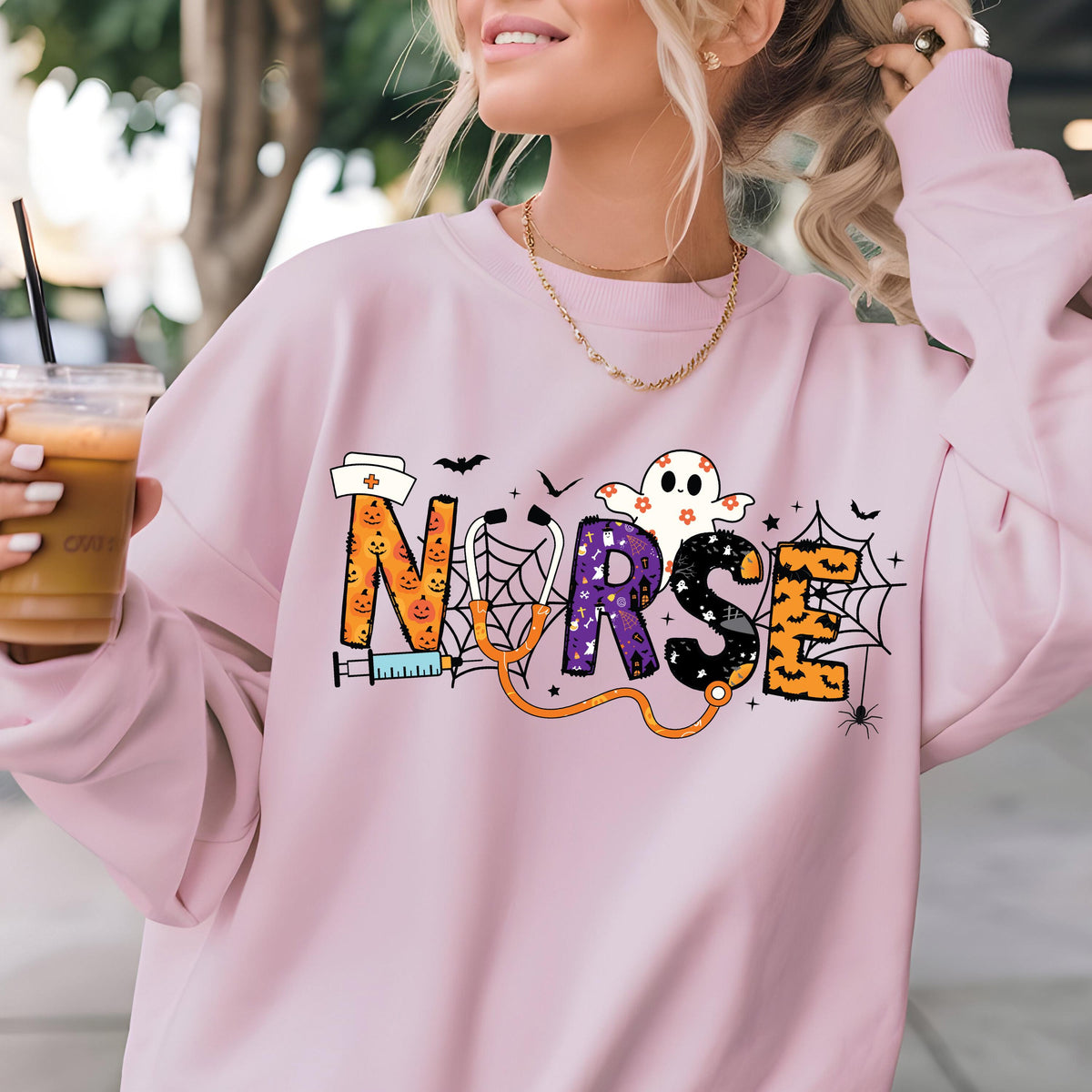 May include: A light pink sweatshirt with a Halloween-themed design. The word "NURSE" is spelled out with orange, purple, and black letters, featuring a ghost, bats, and spiderwebs. A stethoscope and syringe are also included in the design.