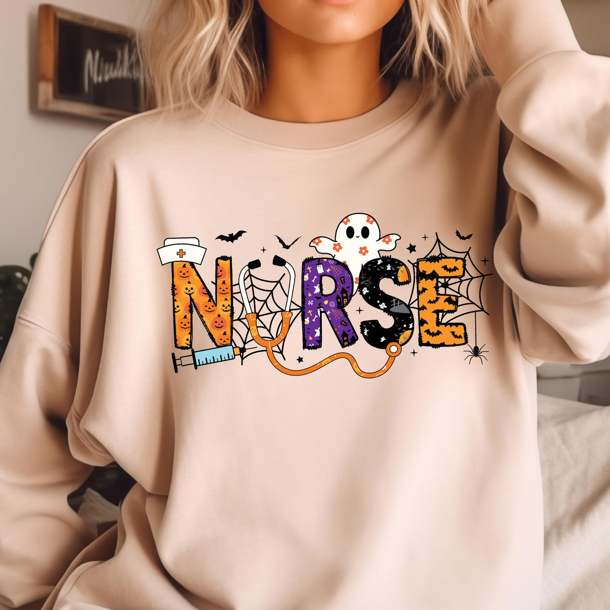 May include: Beige sweatshirt with the word "NURSE" in a Halloween-themed design. The letters are decorated with pumpkins, a ghost, spiderwebs, and a stethoscope. A nurse's hat and syringe are also included in the design.