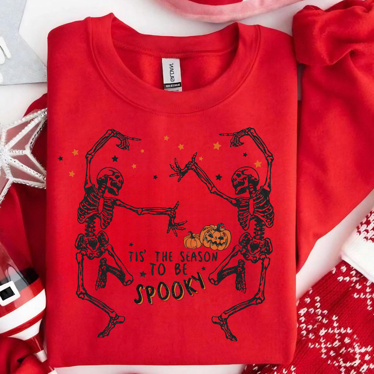May include: Red sweatshirt featuring a black skeleton design with pumpkins and stars. The text "Tis' the season to be spooky" is printed below the skeletons. A festive Halloween-themed apparel item.