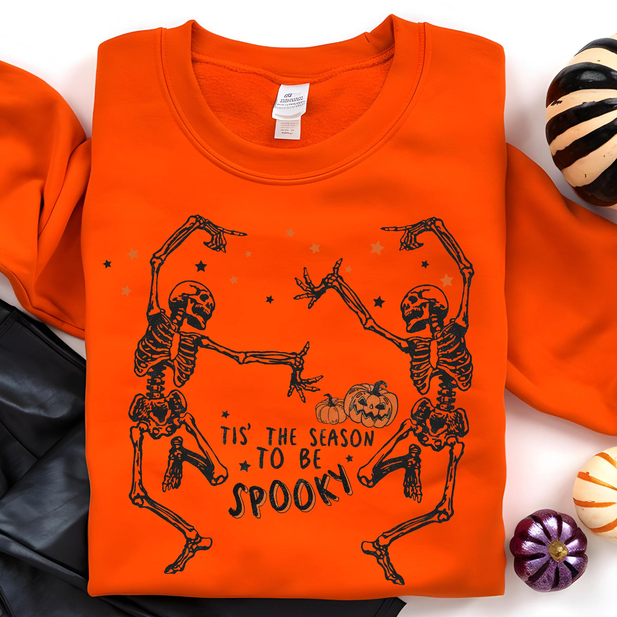 May include: Orange sweatshirt with a black graphic of two dancing skeletons, pumpkins, and stars. The text "Tis' the season to be spooky" is printed below the skeletons. A festive Halloween-themed apparel item.