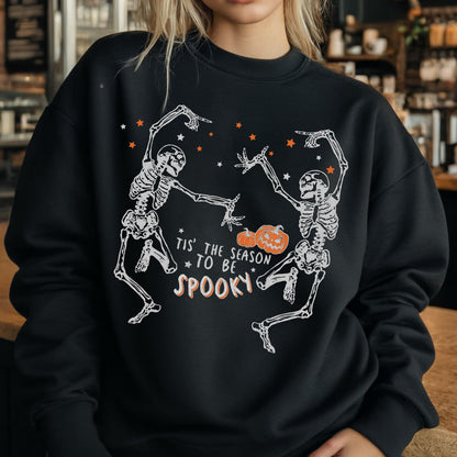May include: Black sweatshirt featuring a white skeleton design with orange stars and pumpkins. The text "Tis' the season to be spooky" is printed below the skeletons. The sweatshirt is a crew neck style.