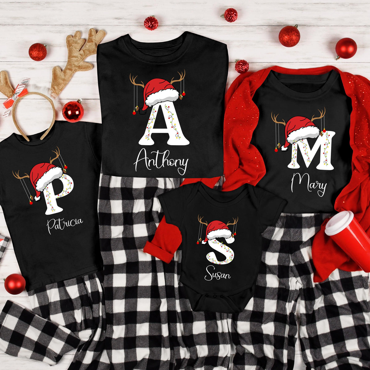 May include: A collection of black t-shirts and a baby bodysuit featuring white letters topped with red Santa hats and reindeer antlers. Each garment displays a name in white script: Anthony, Mary, Patricia, and Susan. The set includes black and white plaid pajama pants and a red cardigan.