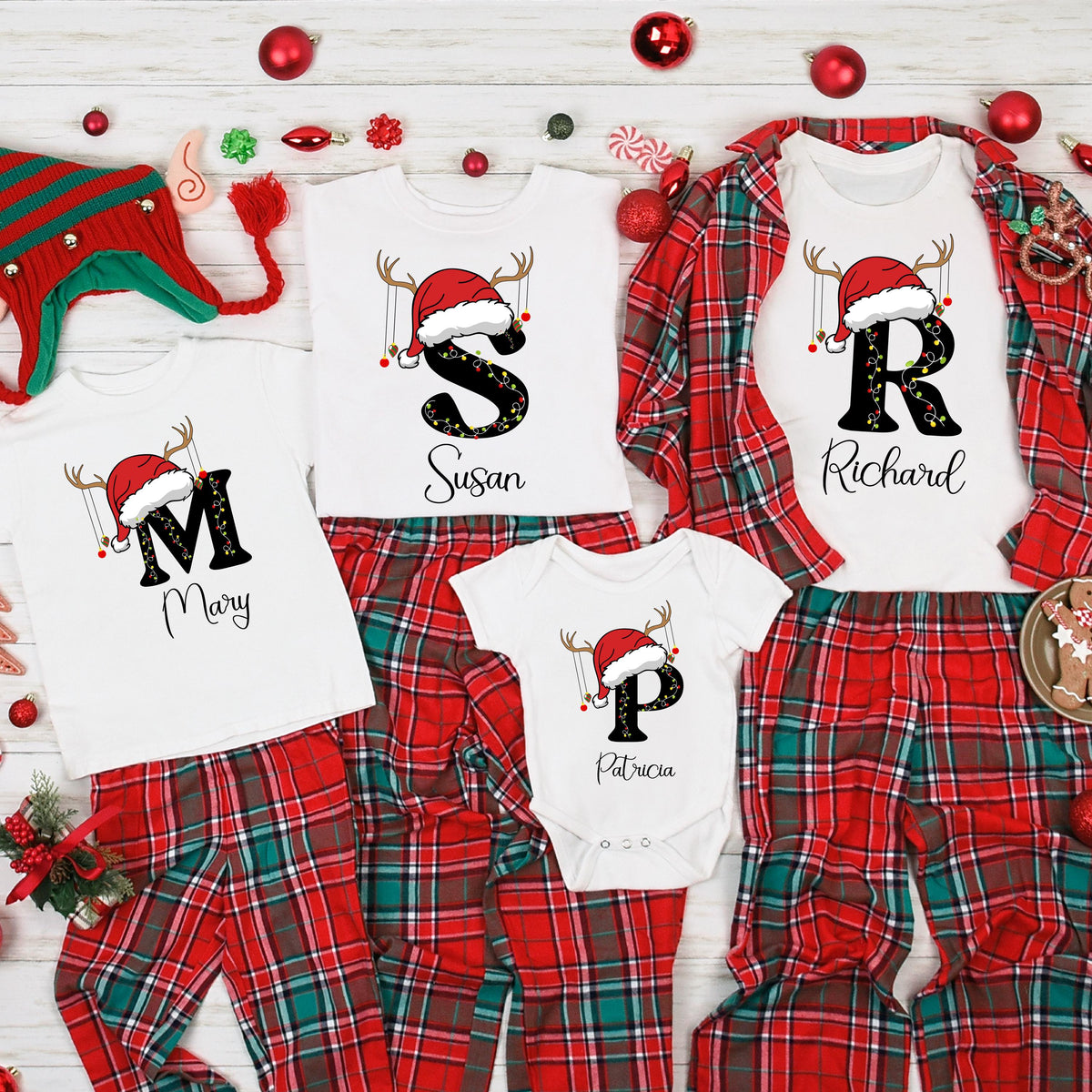 May include: A collection of white t-shirts and a baby bodysuit featuring large black letters M, S, R, and P, each topped with a red Santa hat and reindeer antlers. The names Mary, Susan, Richard, and Patricia are printed below the letters.