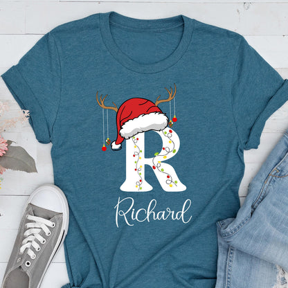 May include: A teal short-sleeved t-shirt featuring a large white letter R decorated with colorful Christmas lights. A red Santa hat with brown reindeer antlers and hanging ornaments sits atop the letter. The name Richard is printed below.