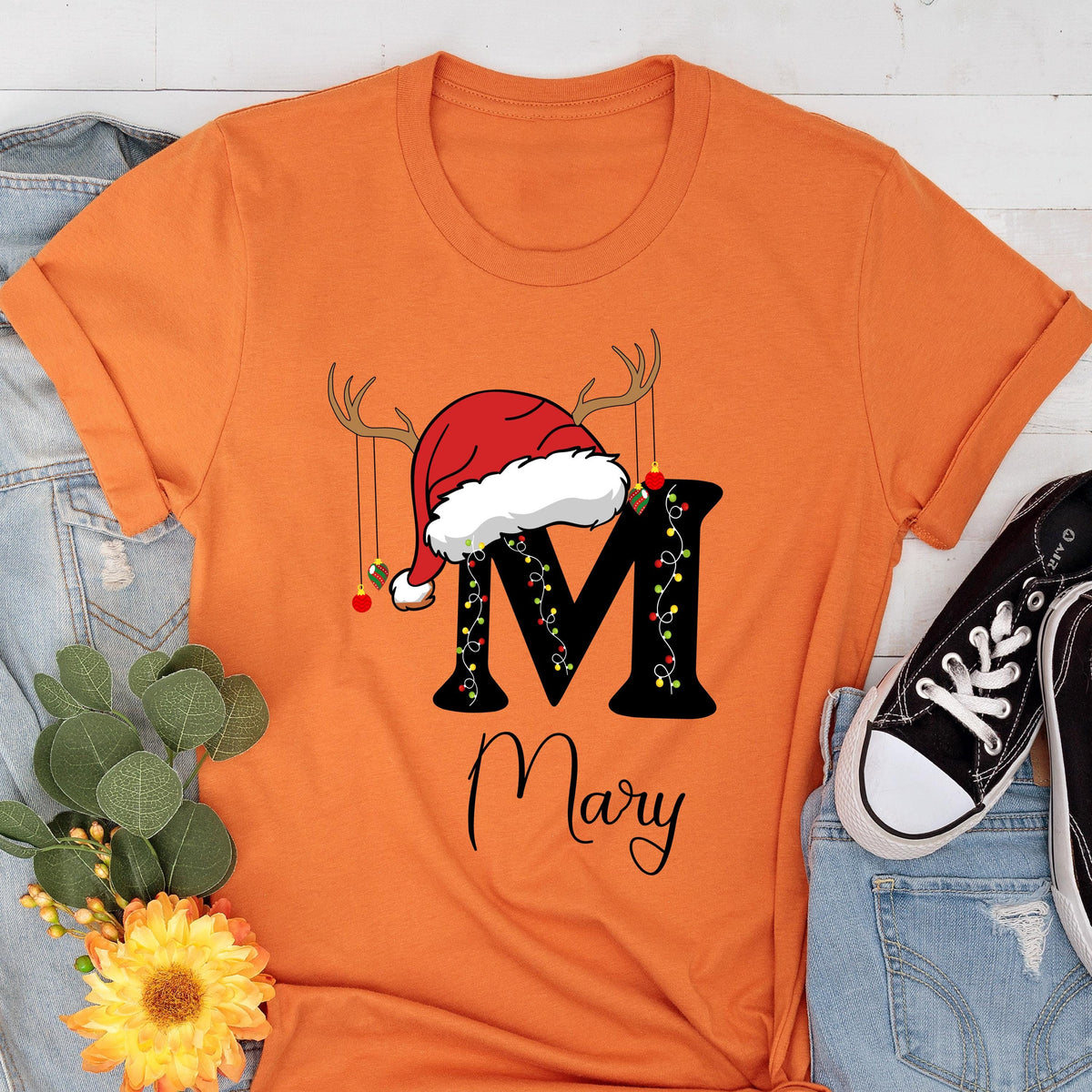 May include: An orange short-sleeved t-shirt featuring a large black letter M decorated with colorful Christmas lights. A red and white Santa hat with reindeer antlers sits atop the letter M. The name Mary is printed below in black script.