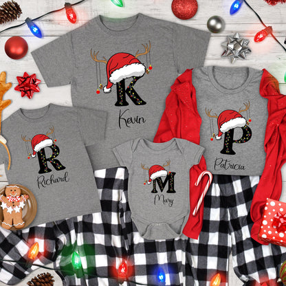 May include: A collection of gray t-shirts and a baby bodysuit featuring large black letters K, P, R, and M. Each letter is topped with a red Santa hat and reindeer antlers, decorated with string lights and ornaments. Names Kevin, Patricia, Richard, and Mary are printed below each letter.