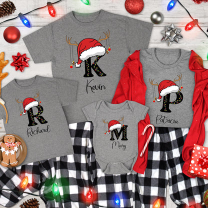 May include: A collection of gray t-shirts and a baby bodysuit featuring large black letters K, P, R, and M. Each letter is topped with a red Santa hat and reindeer antlers, decorated with string lights and ornaments. Names Kevin, Patricia, Richard, and Mary are printed below each letter.