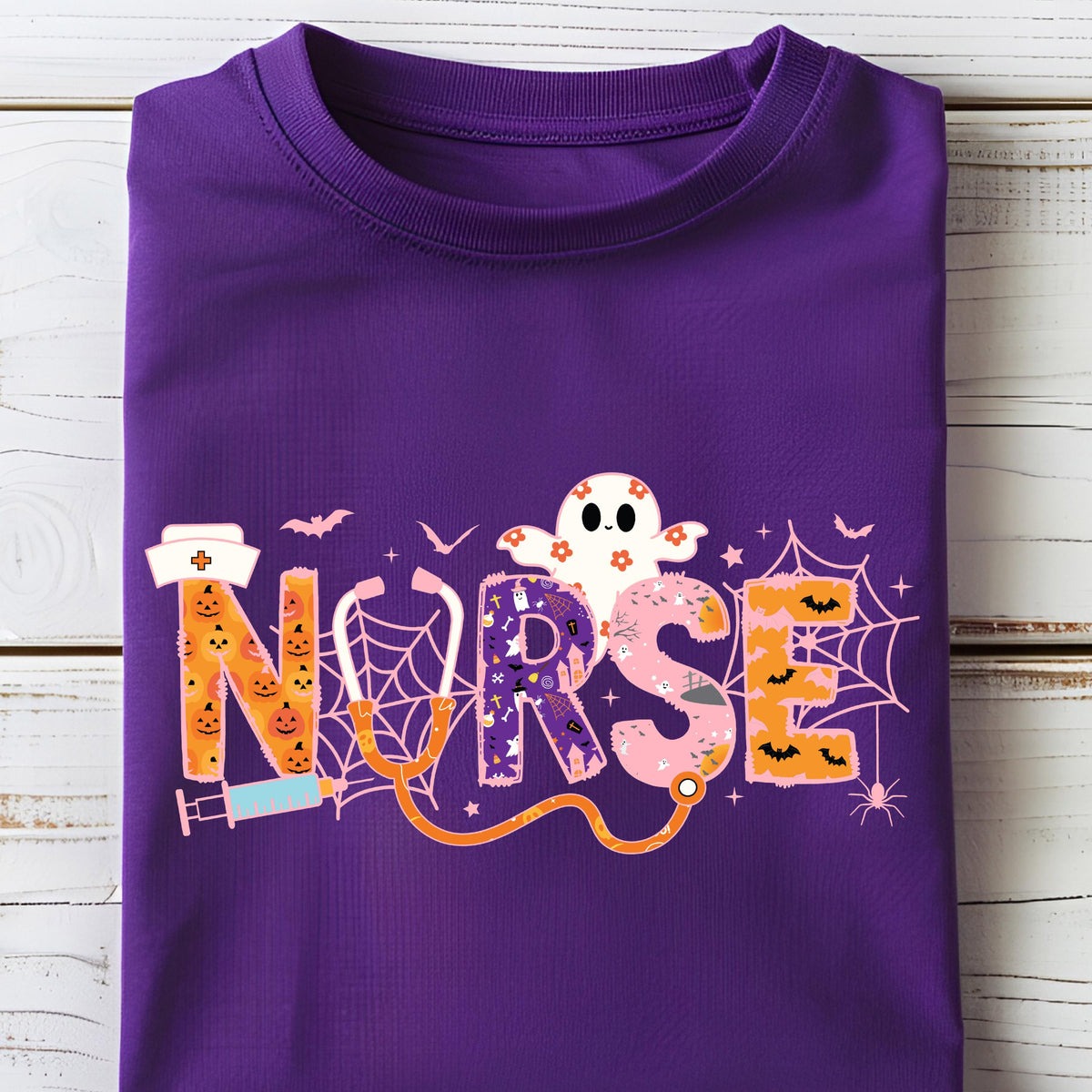 May include: A folded purple t-shirt featuring the word "NURSE" in a Halloween-themed design. The letters are decorated with pumpkins, a ghost, spiderwebs, and bats. A stethoscope graphic is also included.
