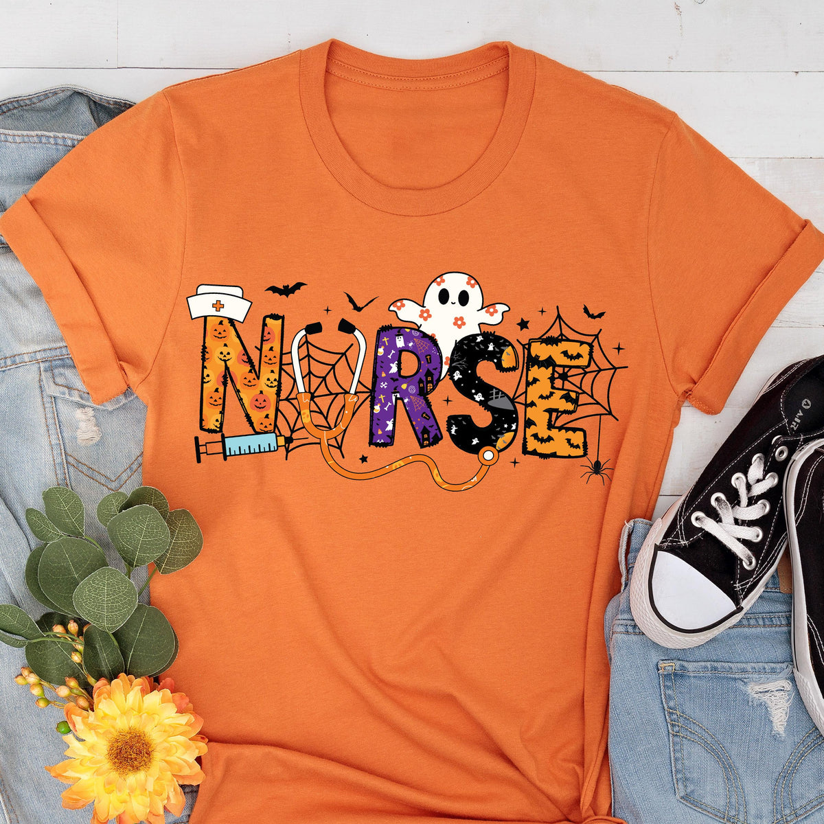 May include: Orange t-shirt with the word "NURSE" in a Halloween-themed design. The letters are decorated with pumpkins, spiderwebs, and a ghost. A stethoscope and syringe are also included in the design. Black sneakers and blue jeans are visible.
